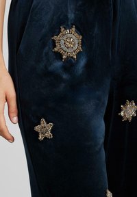 Pantalones de terciopelo marino con insignias decoradas, una con acentos dorados en forma circular y otra con forma de estrella con detalles de piedras preciosas, cerca de una mano.