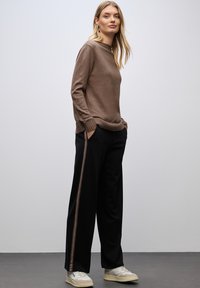 Brauner Pullover mit gerippten Bündchen, kombiniert mit schwarzen, weit geschnittenen Hosen mit einem braunen Streifen. Die Schuhe sind hell gefärbt und haben ein strukturiertes Muster.