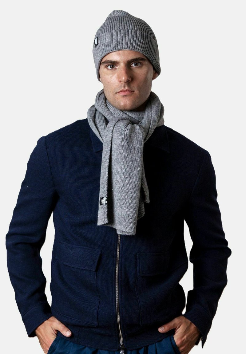Mann mit einer grauen Strickmütze und passendem Schal, navy Farbene Jacke mit Reißverschluss, der mit den Händen in den Taschen vor einem weißen Hintergrund steht.