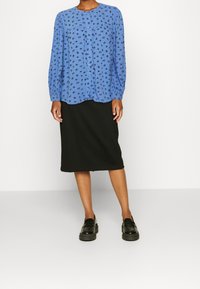 Blaue Bluse mit langen Ärmeln und Blumenmuster, Knopfleiste vorne und Rundhalsausschnitt, kombiniert mit einem schwarzen knielangen Rock und schwarzen Slippern.