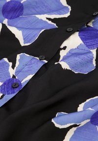 Zwarte stof met grote blauwe bloemenprint en witte accenten, met verschillende zwarte knopen aan de voorkant. Gladde textuur.