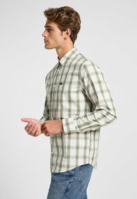 Męska koszula w kratę z długim rękawem w zielono-białym kolorze, z kołnierzykiem zapinanym na guziki, jedną kieszenią na piersi i slim fit. Wykonana z bawełny, o gładkiej fakturze.