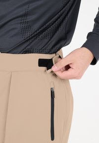 Beige-housut, joissa on sileä pinta, vyötäröllä tarranauha ja sivussa vetoketjullinen tasku. Kangas näyttää kevyeltä.