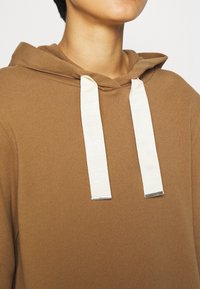 Felpa marrone con cappuccio e tessitura morbida. Presenta coulisse bianche con punte in metallo argentato. Design semplice, senza motivi aggiuntivi.