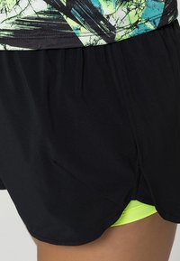 Pantalones cortos deportivos negros con un ajuste holgado y dobladillo curvado, que presentan una capa interna de color verde neón. La tela es suave con un ligero brillo.