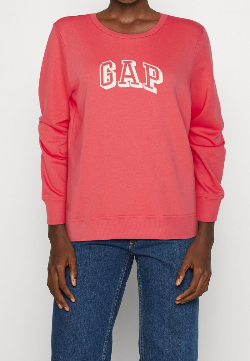 Persona vestida con un sudadera de manga larga color coral de GAP y jeans azules, de pie contra un fondo claro y liso.