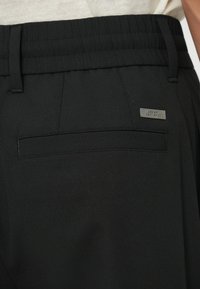 Black tailored pants with a textured, elastic waistband. Features a back pocket with a metal logo accent. Material appears smooth and lightweight.