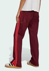 Bordeauxfarbene Sweatpants mit roten Seitenstreifen, lockere Passform, elastischer Bund und akzentuierte Taschen. Kombiniert mit weißen Sneakers mit schwarzen Streifen.