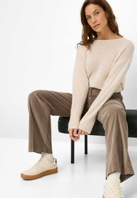 Lichtbeige gebreide trui, ontspannen pasvorm met verlengde mouwen; bruine corduroy broek; off-white schoenen met een gumrubberen zool; zittend op een groene bank.