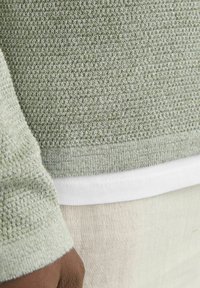 Pull en tricot vert clair avec un motif texturé, des poignets et un ourlet côtelés. Superposé à une chemise blanche avec une texture de tissu lisse.
