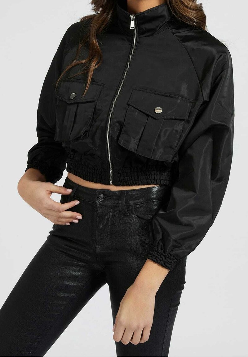 Mujer con una chaqueta negra corta con bolsillos frontales y cremallera, combinada con pantalones negros brillantes de cintura alta, contra un fondo plano.
