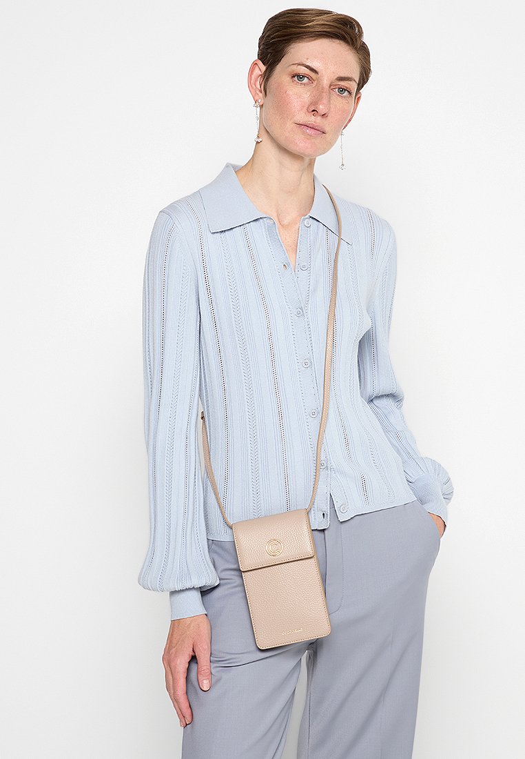 Beige strukturierte Umhängetasche für das Handy mit Klappe und Logo-Akzent, getragen über einem hellblauen Langarmshirt unter einem geknöpften Cardigan.