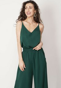 Zielony kombinezon z lekkiego materiału, z dekoltem w serek, cienkimi ramiączkami i elastycznym paskiem w talii; szerokie nogawki; gładka faktura; minimalistyczny design.