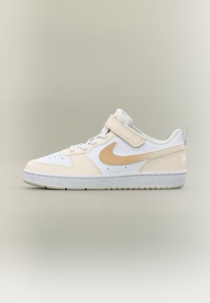 Baskets basses blanches et beiges avec un swoosh Nike marron clair, des lacets et une sangle en Velcro beige sur fond beige.