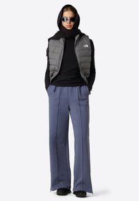 Henkilö, jolla on musta neulepaita, harmaa toppaliivi, siniset leveälahkeiset housut, musta huivi päässä ja valkoiset kehykset aurinkolasit, seisoo kädet taskuissa.