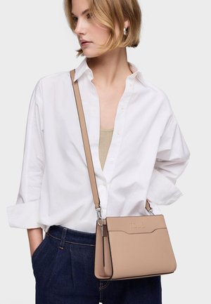 Frau mit kurzen blonden Haaren, die ein weißes Hemd und dunkle Jeans trägt, während sie eine beigefarbene Umhängetasche mit dem eingeprägten Schriftzug "TOUS" auf der Vorderseite trägt.