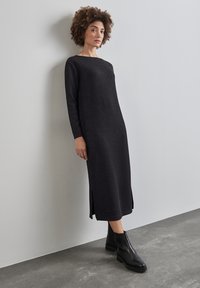 Robe noire longueur cheville avec une silhouette droite, manches longues, décolleté bateau et fentes latérales. Portée avec des bottes noires à la cheville.