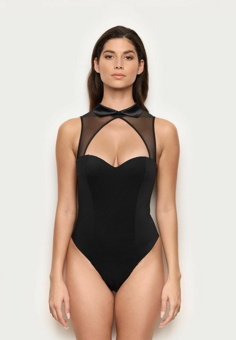 Kobieta w czarnym bezrękawnikowym kombinezonie z przezroczystą górną częścią i satynowym kołnierzykiem, stojąca na jednolitym jasnym tle.