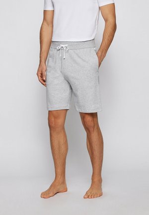 Homme portant un short gris clair à cordon de serrage et un t-shirt blanc, debout pieds nus avec une main dans la poche sur fond uni.