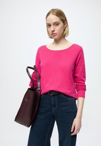 Roza majica z dolgimi rokavi in širokim izrezom, združena s temno modrimi kavbojkami. Model drži temno vinsko rjavo usnjeno torbico z dvema trakovoma.