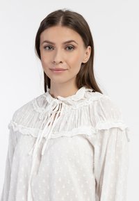 Biała bluzka z falbanistym kołnierzykiem, z fakturowanej tkaniny w kropki, z zapięciem na sznurek. Długie rękawy z marszczeniami.