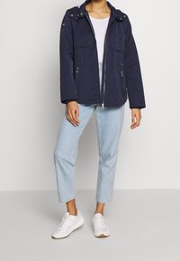 Marineblaue leichte Jacke mit Kapuze, durchgehendem Reißverschluss und zwei Brusttaschen; kombiniert mit hellblauen Jeans und weißen Sneakers.