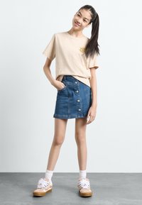 T-shirt din bumbac bej cu un grafic mic galben, combinat cu o fustă din denim albastru cu nasturi în față și buzunare laterale. Adidași maronii completează ținuta.