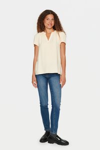 Top corto beige chiaro a maniche corte con scollo a V e una sottile trama verticale, abbinato a jeans blu aderenti e scarpe nere.