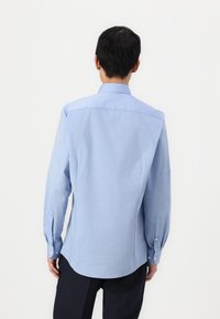 Cămașă albastră deschis, cu mânecă lungă și guler drept, cu manșete, având o textură netedă și un tiv ușor rotunjit.