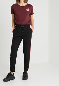 Camiseta de algodón color marrón con el texto "Besa al mundo" y un gráfico de una rosa, acompañada de unos joggers negros con franjas laterales rojas y cintura elástica.