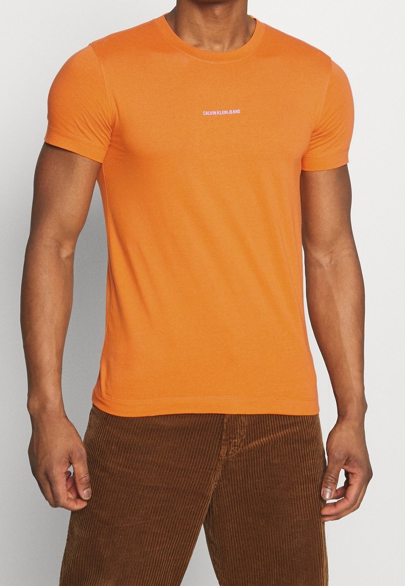 Oranžové bavlněné tričko s kulatým výstřihem a krátkými rukávy, s bílým logem "CALVIN KLEIN JEANS" na hrudi.