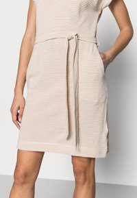 Robe rayée beige clair avec des manches courtes, une ceinture à nouer à la taille et des poches latérales. Texture douce et design arrivant au genou.