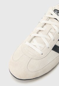 Ανοιχτό μπεζ σουέτ αθλητικό παπούτσι με λευκά κορδόνια, μαύρες λεπτομέρειες και στρογγυλερή μύτη. Διακριτικό ραφικό μοτίβο κατά μήκος του πάνω μέρους και της σόλας.