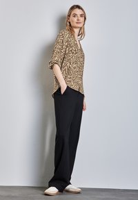 Blusa marrone a fantasia con scollo a V e maniche arrotolate, abbinata a pantaloni neri a gamba larga e sneakers chiare. Tessuto morbido, vestibilità rilassata.