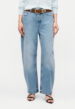 Žena oblečená v svetlomodrých džínsach s rozšírenými nohavicami a hnedým opaskom, v kombinácii s čiernymi sandálmi na podpätku so šnúrkami.