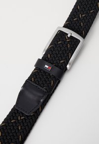 Geweven zwarte riem met zilveren metalen gesp, heeft bruin stiksel, leren accenten en een klein meerkleurig logo-detail.