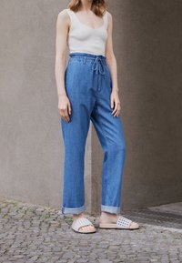 Top a costine beige leggero, pantaloni in denim blu larghi con cintura da annodare e sandali bianchi intrecciati con suola naturale.