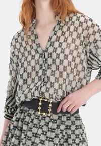 Genomskinlig blus med en geometrisk mönstring i brunt, gräddvitt och svart. Har en V-hals, långa ärmar och lager på lager-texturer. Dekorativt bälte med guldfärgad accenter.