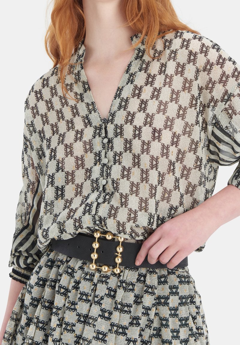 Genomskinlig blus med en geometrisk mönstring i brunt, gräddvitt och svart. Har en V-hals, långa ärmar och lager på lager-texturer. Dekorativt bälte med guldfärgad accenter.