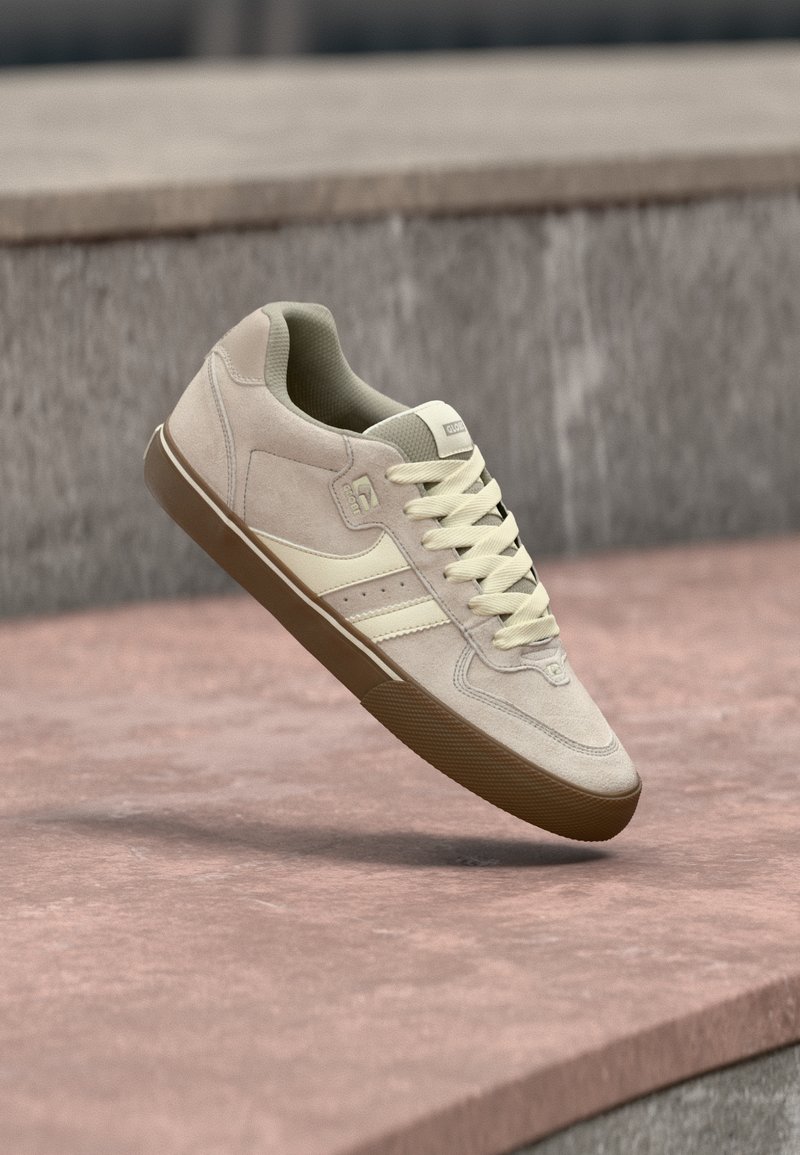 Baskets en daim beige avec des accents jaune clair, présentant une surface texturée, un bout arrondi et une semelle en gomme en caoutchouc, affichées en suspension dans les airs.