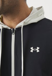 Felpa con zip di colore blu navy, con coulisse e colletto bianchi. Presenta un logo bianco ben visibile sul lato sinistro del petto. Tessuto liscio e leggero.