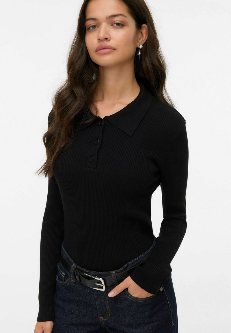Polo noir à manches longues côtelé avec un col et une patte de boutonnage à trois boutons. Porté avec un jean bleu foncé et une ceinture noire.