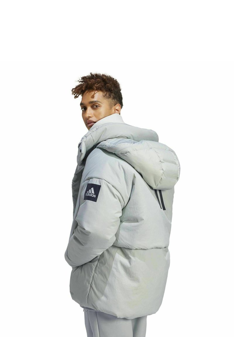 Jasnoszara kurtka puchowa z wysokim kołnierzem, oversize'owym kapturem i czarnym logo Adidas na rękawie. Gładki materiał i pikowana tekstura.