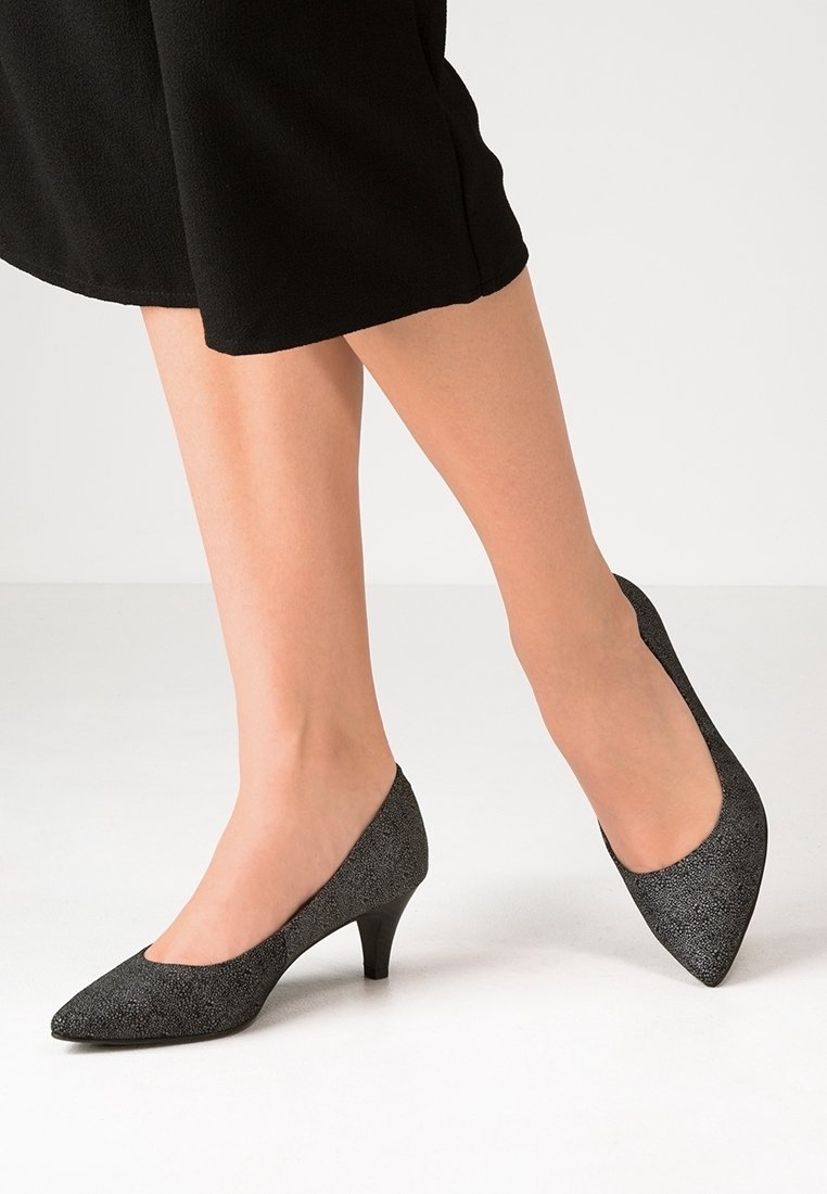 Femme portant des chaussures noires texturées à talons bas et bouts pointus, avec une jupe noire mi-longue, sur un fond uni.