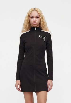 Kobieta z długimi kręconymi blond włosami, ubrana w dopasowaną czarną sukienkę na suwak z białymi paskami na ramionach i logo Puma na piersi.