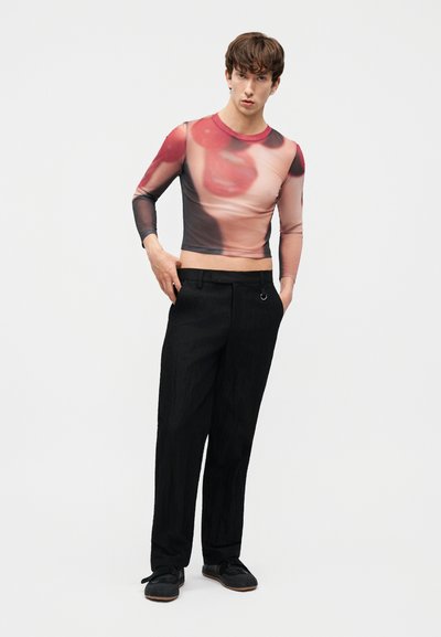 Hombre joven con una camiseta de manga larga, corta, de diseño abstracto en rojo y rosa, pantalones anchos negros y zapatos negros, de pie con las manos en los bolsillos.