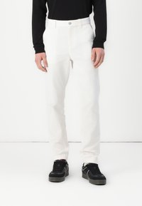 Witte katoenen broek met een rechte pijp, voorzien van een knoopsluiting en zijzakken, gedragen met zwarte sneakers.