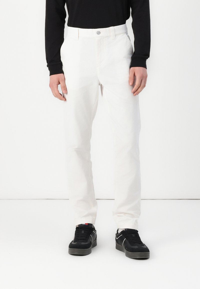 Witte katoenen broek met een rechte pijp, voorzien van een knoopsluiting en zijzakken, gedragen met zwarte sneakers.