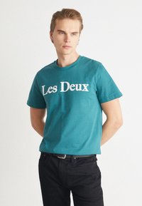 Tirkīza krāsas kokvilnas T-krekls ar baltu tekstu "Les Deux" krūtīs; īsas piedurknes un apaļa kakla izgriezums. Modelis valkā melnus džinsus.