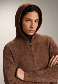 Mladý človek so stredným odtieňom pokožky a kučeravými vlasmi nosí hnedú kapucňu so zipsom, má založené ruky a stojí pred jednoduchým neutrálnym pozadím.